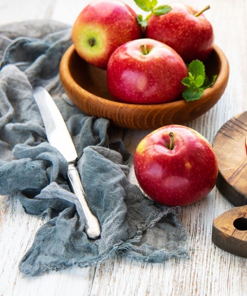 A wooden bowl full of apples and an apple next to a blue napkin and knife