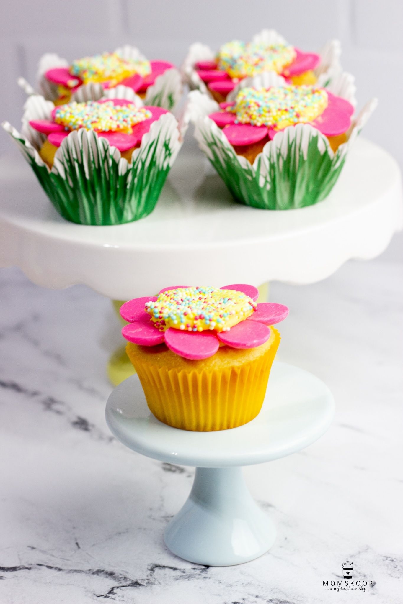 A Daisy cupcake on a light blue cake stand and four Spring cupcakes on a cake stand behind it