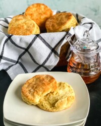 A buttery buttermilk biscuit cut in half on plate next to a basket full of biscuits