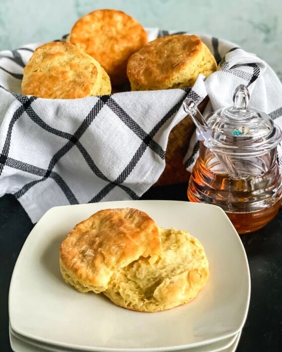 A buttery buttermilk biscuit cut in half on plate next to a basket full of biscuits