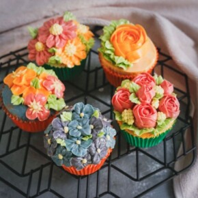 Five cupcakes decorated with a variety of flowers made from buttercream sitting on a black wired rack