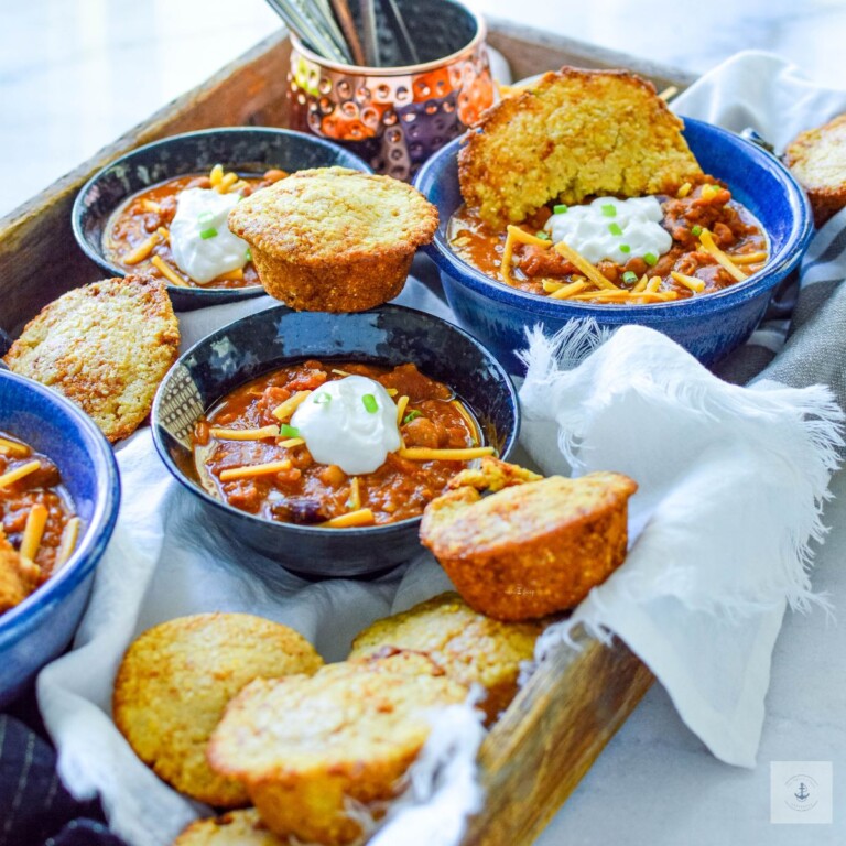 Blue bowl full of Chili with Kielbasa sausage, half of a cornbread muffin, a dollop of cream cheese, diced green onions and cheese.