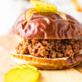 Homemade sloppy joes on a pretzel bun with pickle slices on a wooden board