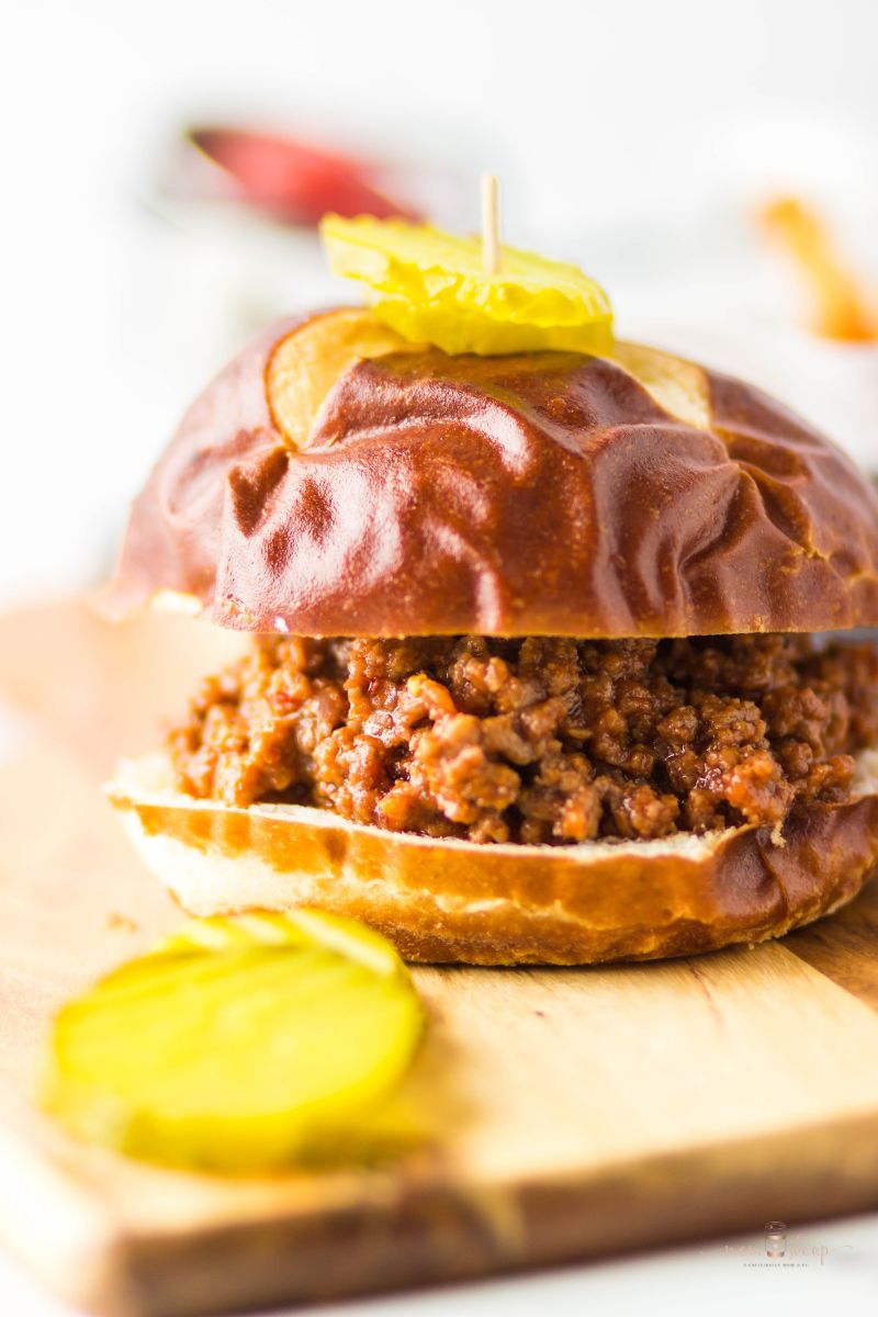 Homemade sloppy joes on a pretzel bun with pickle slices on a wooden board