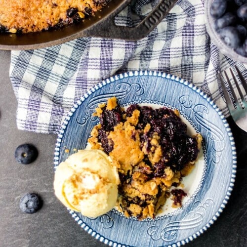Blueberry crumble with a scoop of ice cream on a blue and white plate with a gingham blue and white napkin.
