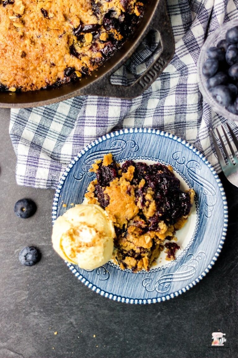 Blueberry crumble with a scoop of ice cream on a blue and white plate with a gingham blue and white napkin.
